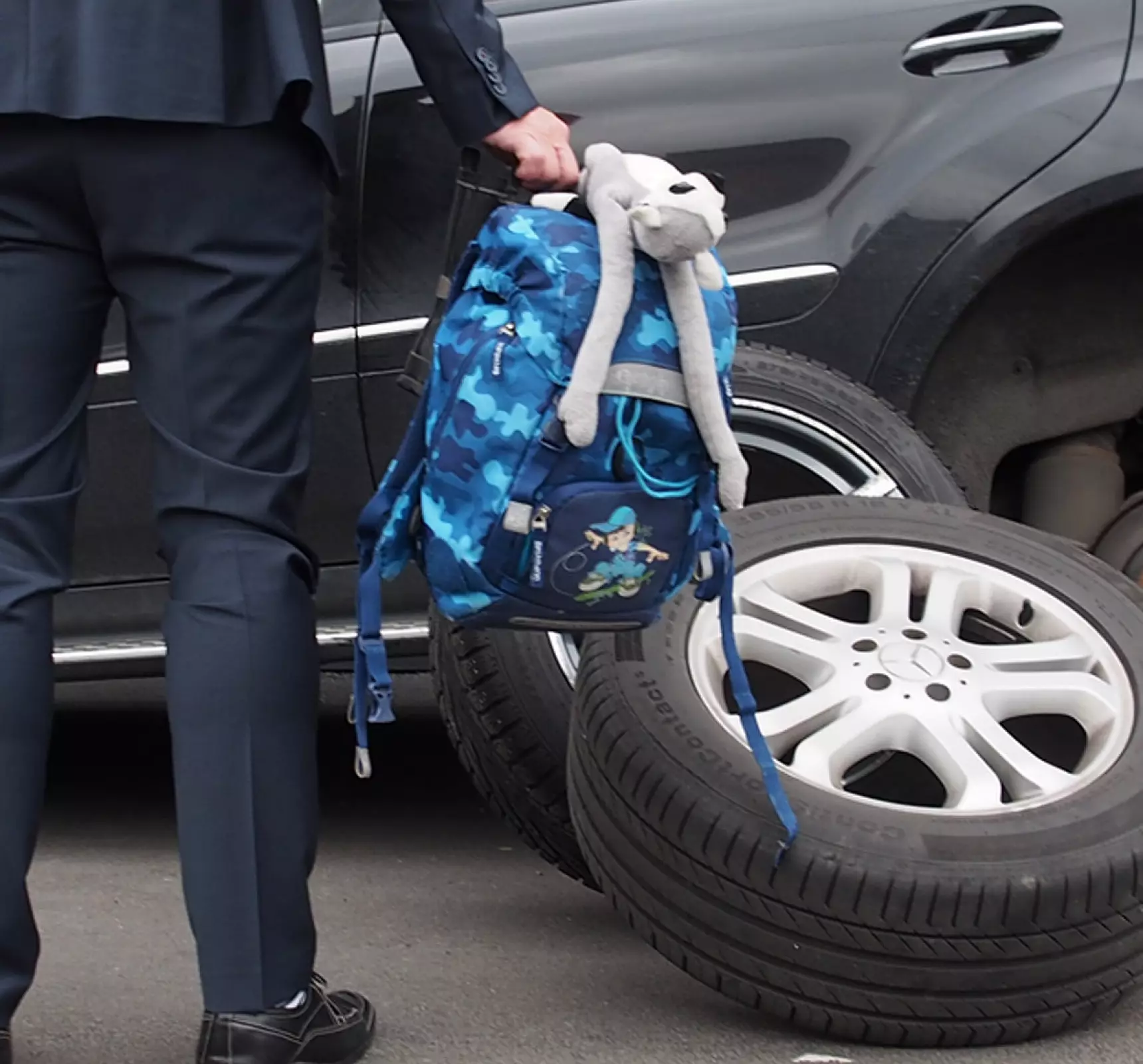 Mann med dress som holder en ransel med bamse foran en bil som er klar for dekkskifte